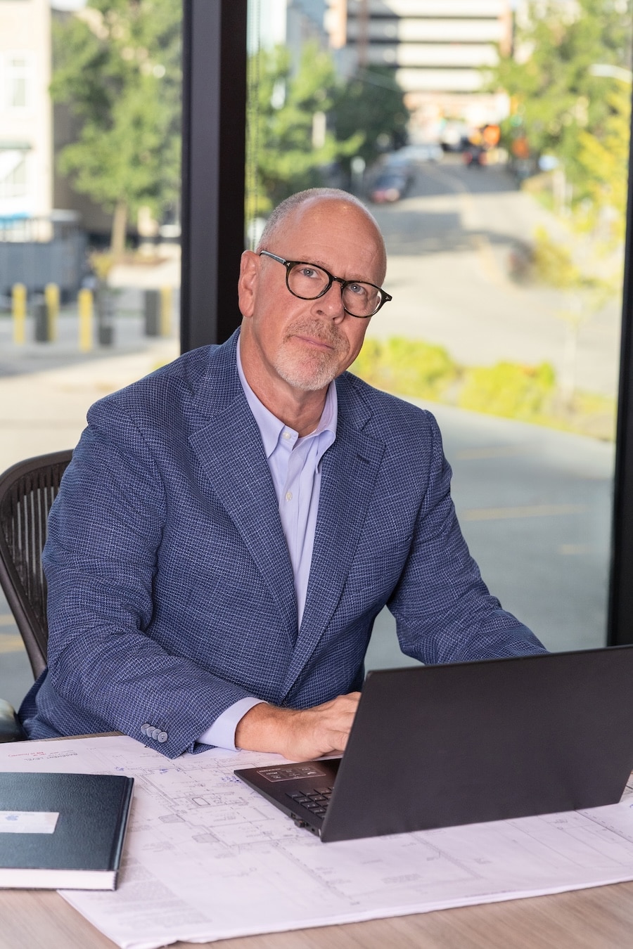 Bald man in glasses and blue blazer sits at a desk with a laptop and papers, looking at the camera.